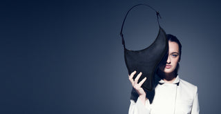 A model in a white suit holds up the Arcana black bag against a dark blue studio background.